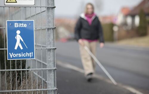 Eine Fußgängerin ist verschwommen im Hintergrund zu sehen. Sie spaziert mit ihrem Langstock auf dem Gehweg. An einem Gitterzaun im Vordergrund hängt ein Schild mit der Aufschrift „Bitte Vorsicht!“ und einem Stockgänger-Symbol.