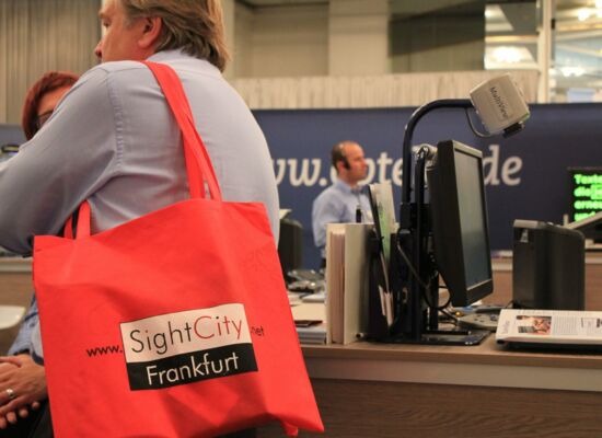 Ein Bild aus dem Messegeschehen. Im Vordergrund leuchtet eine rote Tasche, auf der "SightCity Frankfurt" steht.