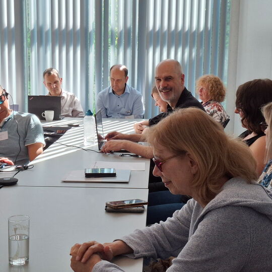 Beim Workshop „Überregionale Kooperation“: Teilnehmerinnen und Teilnehmer sitzen an Tischen. Vor manchen liegt ein Smartphone, andere haben ihr Laptop aufgeklappt. Auch Workshop-Leiter Reiner Delgado bedient ein Laptop, während er lächelnd Richtung Kamera