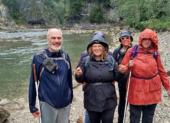 Reiner Delgado (links) und vier weitere Personen der Wandergruppe stehen lachend an einem steinigen Ufer. Sie tragen Outdoor-Jacken, Rucksäcke, Wanderschuhe und teilweise einen Langstock.
