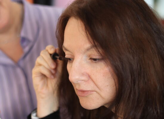 Mit einem Wimpernbürstchen trägt Ute Stephanie Mansion Farbe auf die Wimpern des rechten Auges auf. Sie hat mittellanges dunkles Haar und blickt konzentriert.