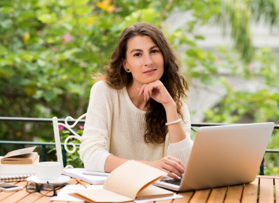 Eine Frau mit langen braunen Haaren sitzt draußen an einem Tisch. Im Hintergrund sind verschwommen Bäume zu sehen. Vor ihr liegen mehrere Bücher und ein Laptop. Die Hand unter dem Kinn haltend, blickt sie versonnen in die Kamera.