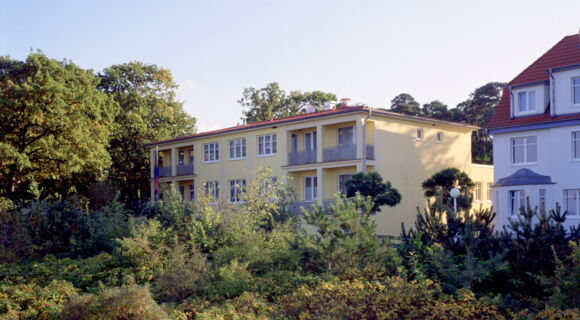 Das AURA-Hotel Boltenhagen, ein gelbes eckiges Gebäude mit schlichten Balkonen und rotem flachen Dach, steht zwischen Büschen und Bäumen. Daneben ist ein weiteres großes, weißes Haus zu sehen.