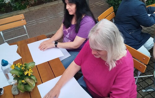 Zwei Frauen sitzen an einem Holztisch und studieren mit den Fingern je eine Wanderkarte in Braille. Die Frau links ist Yvonne Bühr. Sie hat langes dunkles Haar.