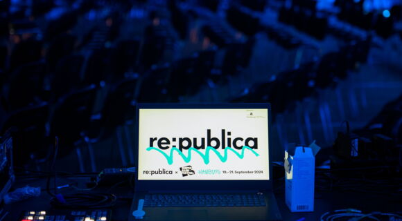 Ein Laptop auf einem Technik-Tisch zeigt das Logo der „re:publica". Im Hintergrund sind viele leere Stuhlreihen in einem blauen Licht beleuchteten Raum zu sehen. Kabel und Technikgeräte liegen auf dem Tisc