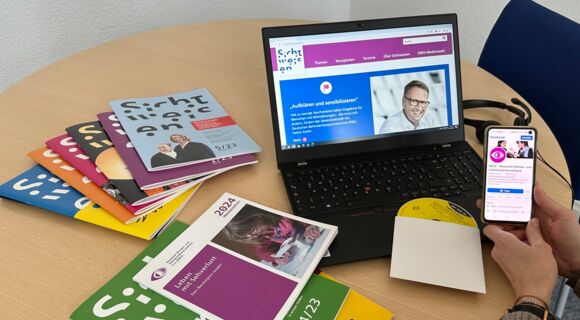 Auf einem Tisch steht ein Laptop, das eine Seite von Sichtweisen-online zeigt. Daneben ein Stapel Sichtweisen-Hefte, die Publikation „Weitersehen 2024“ und eine Daisy-CD.