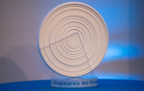 Der Hörspielpreis der Kriegsblinden, eine weiße runde Scheibe auf einem durchsichtigen Sockel, steht auf einem blauen Untergrund. Der runde Pokal erinnert durch ringförmig angeordneten Rillen an eine Musikplatte.