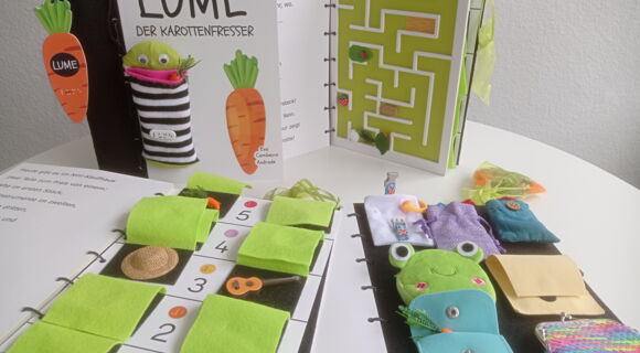 Interaktives Tastbuch „Lume der Karottenfresser“ mit Stofffiguren, Taschen, Labyrinth und verschiedenen taktilen Spielelementen auf mehreren geöffneten Buchseiten.