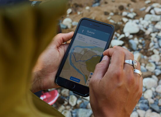 Jemand steht draußen auf Steinen und hält ein Handy mit der Navigations-App Komoot in der Hand. Darauf ist eine Route zu sehen. Der Mensch hat silberne Ringe und trägt eine grüne Jacke.