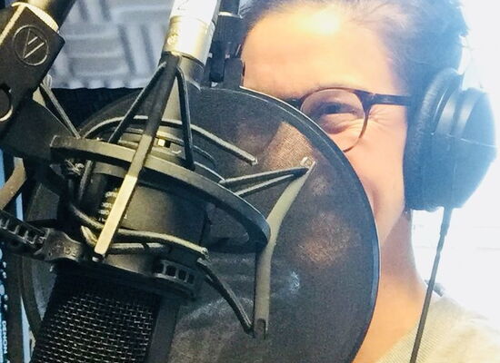 Melanie Wölwer spricht in eine große Aufnahmeanlage, von der sie halb verdeckt wird. Ihre dunklen Haare hat sie nach hinten gebunden, sie trägt eine dunkel umrandete Brille.