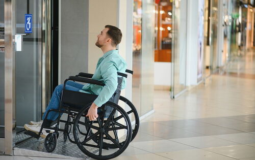 Ein Rollstuhlfahrer vor einer Tür in einem Einkaufszentrum. Auf der Glastür ist ein Aufkleber mit Rollstuhl angebracht.