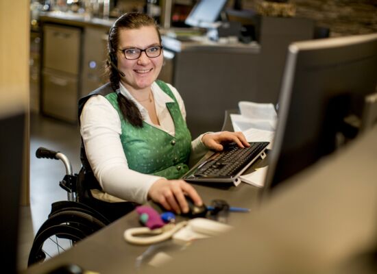 Eine Frau mit breitem Lächeln sitzt in einem Büro am Computer. Sie nutzt einen Rollstuhl. Sie hat eine grüne Weste mit Knöpfen an, trägt eine Brille und hat ein Headset auf dem Kopf.