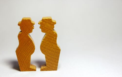 Auf einem weißen Hintergrund stehen sich zwei kleine Holzfiguren gegenüber. Sie sehen aus wie zwei untersetzte Männer mit Hut.