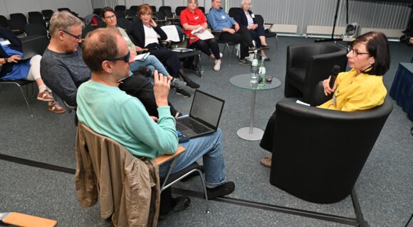 Workshop in einem größeren Raum: Die Teilnehmerinnen und Teilnehmer sitzen in einer Reihe; vorne befinden sich ein Mann und eine Frau, die jeweils ein Mikro in der Hand halten.