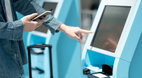 Jemand steht vor einem Automaten und zeigt mit dem Finger darauf. In der anderen Hand hält er ein Handy und ein kleines Heft.