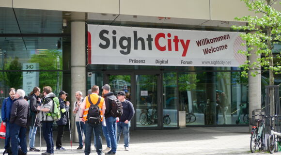 : Vor dem Eingang der Sightcity steht eine Gruppe von Menschen. Viele von Ihnen haben einen Langstock dabei. Sie tragen Rucksäcke. Über der Tür hängt ein Willkommens-Banner.