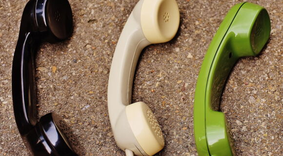 Drei klassische Telefonhörer in Schwarz, Beige und Grün liegen nebeneinander auf einem Korkuntergrund.