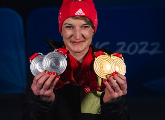 Anna-Lena Forster hält in beiden Händen vier Medaillen hoch. Sie trägt eine rote Mütze und lächelt.