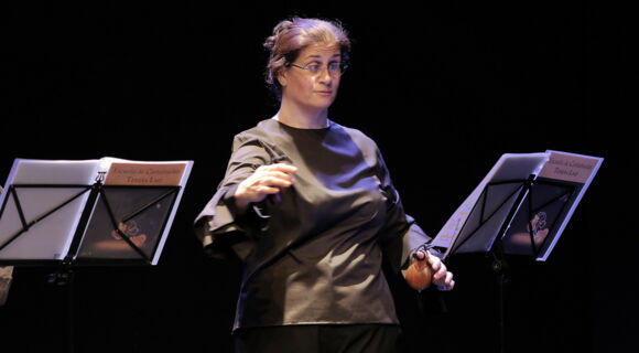 Bárbara Martín Muñoz steht auf einer Bühne, mit beiden Händen spielt sie je eine Kastagnette. Sie blickt auf Noten auf einem Notenständer. Ihr Haar hat sie hochgebunden, sie trägt eine Brille.