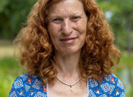 Brigitte Buchsein hat langes lockiges helles Haar. Sie trägt ein Shirt mit Blumen und eine Kette mit einem kleinen Kreuzanhänger.