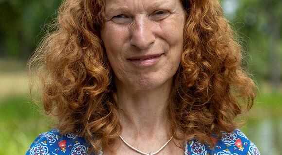 Brigitte Buchsein hat langes lockiges helles Haar. Sie trägt ein Shirt mit Blumen und eine Kette mit einem kleinen Kreuzanhänger.