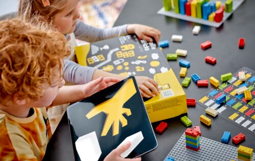 Zwei Kinder sitzen an einem Tisch. Vor sich liegt ein Karton mit Braille-Bricks, darum herum Legosteine.