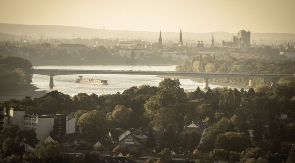 Blick in die Ferne auf die Silhouette von Bonn. Eine lange Brücke spannt sich in der Mitte des Bildes über den Rhein.