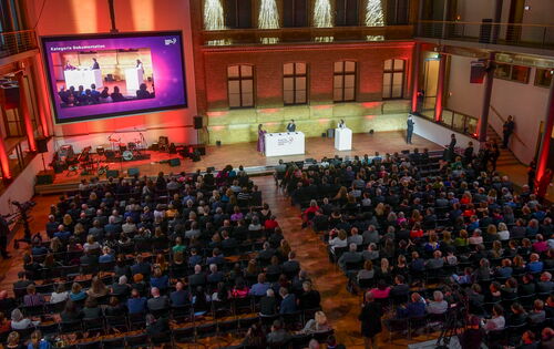 Von schräg hinten wird über das festlich gekleidete Publikum hinweg die Bühne fotografiert. Vorne steht Moderatorin Nadine Heidenreich an einem Pult.