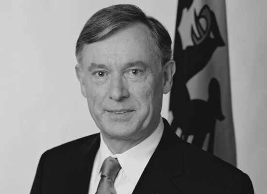 Horst Köhler schaut leicht lächelnd in die Kamera. Er hat sein Haar auf der linken Seite gescheitelt. Er trägt einen schwarzen Anzug und eine helle Krawatte. Im Hintergrund steht eine Fahne mit Reichsadler-Abbildung.