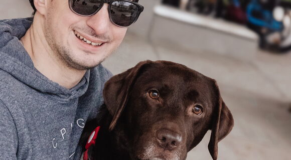 Stephan Dietrich mit seiner Hündin Alma. Er hat kurzes dunkles, leicht lockiges Haar und trägt eine Sonnenbrille.