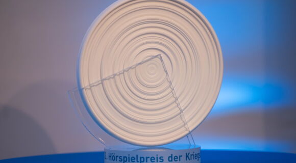 Der Hörspielpreis der Kriegsblinden, eine weiße runde Scheibe auf einem durchsichtigen Sockel, steht auf einem blauen Untergrund. Der runde Pokal erinnert durch ringförmig angeordneten Rillen an eine Musikplatte.