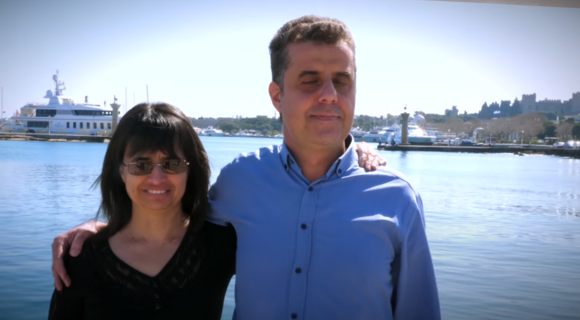 Links steht eine Frau mit mittellangen dunklen Haaren und dunkler Sonnenbrille. Neben ihr ein Mann mit kurzen dunklen Haaren und geschlossenen Augen. Er legt den Arm um sie. Yachten, Wasser und blauer Himmel im Hintergrund.