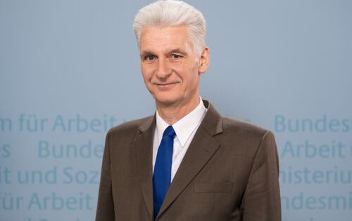 Rolf Schmachtenberg hat weißes, zurückgekämmtes Haar. Er trägt einen braunen Anzug, ein weißes Hemd und eine blaue Krawatte. Im Hintergrund ist unscharf der Text: „Bundesministerium für Arbeit und Soziales“ angedeutet.