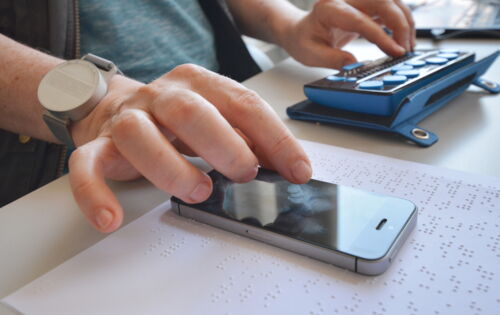 Zwei Hände sind zu sehen: Die linke bedient eine Braillezeile, die rechte ein Smartphone. Das Smartphone liegt auf einer mit Brailleschrift bedruckten Seite.