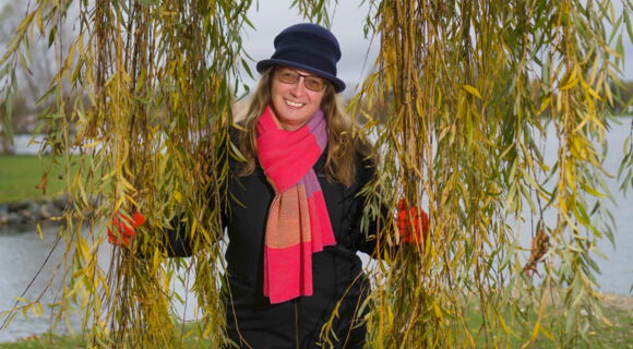 Kathleen Starke, umrahmt von den Ästen und Blättern einer Trauerweide. Sie hat langes helles Haar und trägt eine getönte Brille. Mütze und Jacke sind schwarz, ihr großer Schal ist pink-lila-gestreift.