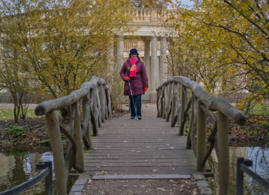 Kathleen Starke geht über eine Holzbrücke, die von hohen Pflanzen umgeben ist. Sie trägt eine lila Jacke, eine schwarze Mütze und einen pink-lila gestreiften Schal.