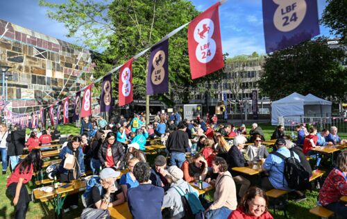 Berliner Platz neben der Liederhalle. Auf einer Rasenfläche stehen im Sonnenlicht Bierbänke und Tische, an denen zahlreiche Menschen sitzen. Über ihnen ist eine Leine gespannt, an der Fähnchen mit dem Logo des Louis Braille Festivals hängen.