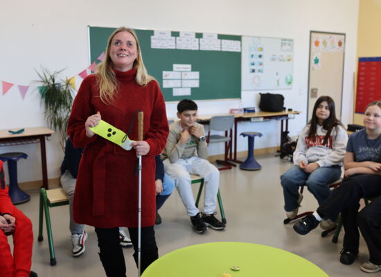Mandy Kugel steht in einer Schulklasse in einem Stuhlkreis; ein Teil der Kinder ist zu sehen. Sie hat lange blonde Haare und trägt einen warmen Pullover. Mit der linken Hand hält sie ihren Langstock, mit beiden Händen zeigt sie eine Armbinde mit drei Punk