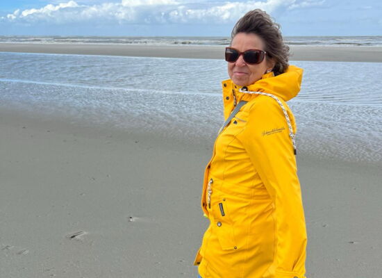 Manuela Dolf geht in gelbe Regenjacke, Jeans und Sneakern an einem Strand entlang. Sie trägt eine Sonnenbrille.