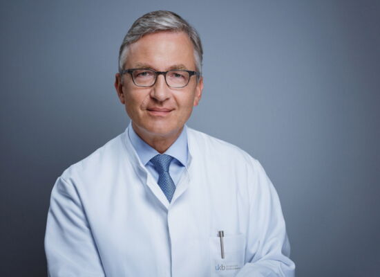 Prof. Dr. Frank G. Holz trägt einen Arztkittel über Hemd und Krawatte. Er hat kurzes, seitlich gescheiteltes graues Haar. Seine Brille ist dunkel und rechteckig.