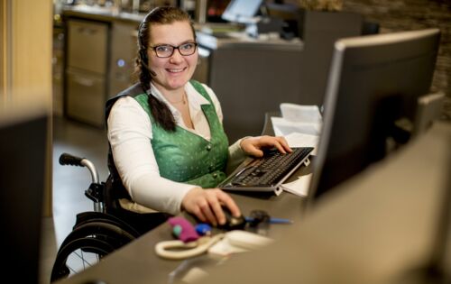 Eine Frau mit breitem Lächeln sitzt in einem Büro am Computer. Sie nutzt einen Rollstuhl. Sie hat eine grüne Weste mit Knöpfen an, trägt eine Brille und hat ein Headset auf dem Kopf.