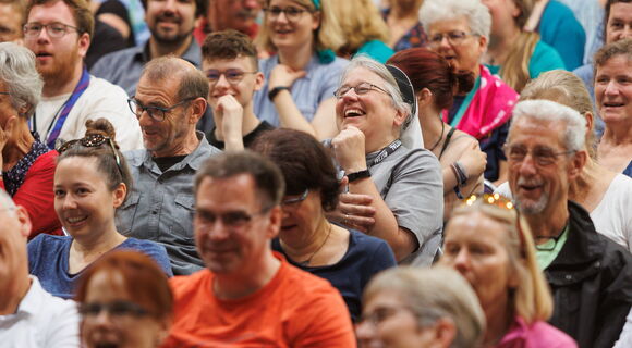 Bild eines Publikums beim Kirchentag 2023 in Nürnberg: Lachende und lächelnde Menschen verschiedenen Alters blicken in eine Richtung. Einige tragen kurzärmelige T-Shirts.