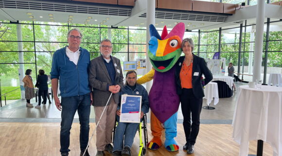 Gruppenbild: Drei Männer, einer davon im Rollstuhl, eine Frau und das Kampagnen-Maskottchen, ein Chamäleon, stehen in einer Empfangshalle. Heinrich Schaudt, Vorsitzender Arbeitsgemeinschaft Barrierefreiheit Rhein-Neckar, präsentiert die Gewinner-Urkunde.