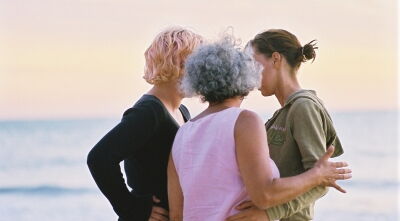 Drei Frauen stehen kreisförmig zusammen und besprechen etwas. Ihre Gesichter sind nicht zu sehen. Zwei von ihnen halten sich gegenseitig unterstützend am Arm. Zwei der Frauen haben nackenlanges blondes lockiges Haar, die dritte hat dunkleres Haar.