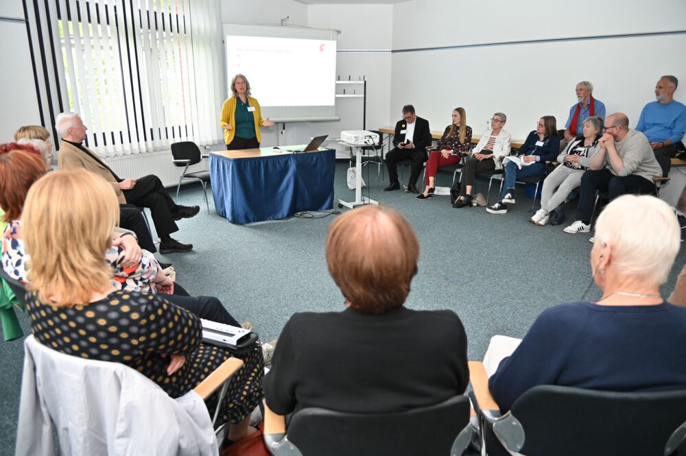 Die Workshopteilnehmenden sitzen in einem großen Kreis beisammen. Eine Frau hält, an einem Tisch mit bodenlanger, blauer Tischdecke stehend, einen Impulsvortrag. Eine Präsentation wird hinter ihr an die Wand geworfen.