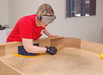 Eine Frau steht an einem Showdown-Tisch mit hohem Rand und Toren. Sie schlägt mit einem flachen, kleinen Schläger den Ball zurück. Ihre Augen sind von einer Brille bedeckt.
