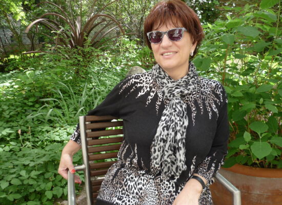 Silja Korn sitzt auf einer Bank, hinter der viele Pflanzen stehen. Sie hat kurzes dunkles Haar und trägt eine Sonnenbrille. Der gezackte Kragen ihres Shirts hat, ebenso wie ihr Tuch, einen Leoprint.