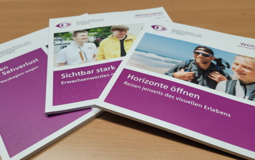 Drei Ausgaben der DBSV-Jahrespublikation "Weitersehen" liegen aufgefächert auf einem Tisch. "Leben mit Sehverlust", "Sichtbar stark" und "Horizonte öffnen" steht weiß auf purpur auf dem jeweiligen Heft.