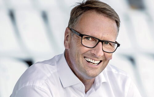 Stefan Kiefer vor verschwommenem Hintergrund: Er hat kurzes, seitlich gescheiteltes Haar, trägt eine dunkle Brille, ein weißes Hemd und lächelt.