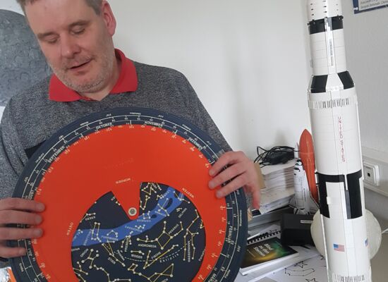 Gerhard Jaworek befühlt eine große runde Sternenkarte, die er in den Händen hält. Neben ihm steht das Modell einer Rakete. Jaworek hat kurzes dunkleres Haar.